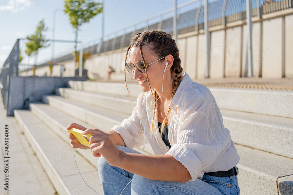 Smiling beautiful woman holding mobile phone listening music, watching video on the street. Happy hipster female using mobile app shopping online outdoors.