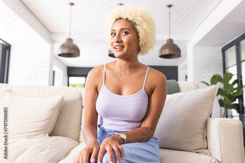 Young biracial woman with blonde curly hair sits on beige sofa, wearing a light blue dress and a sil