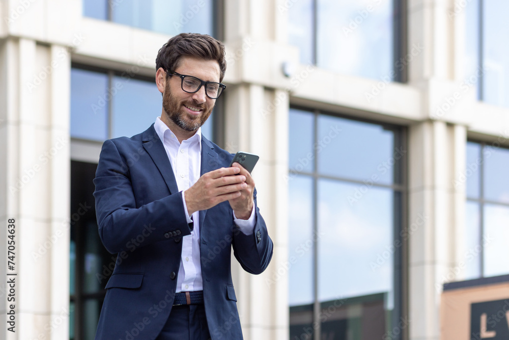 Smiling businessman in suit using smartphone outdoors with office building Smiling businessman in suit using smartphone outdoors with office building