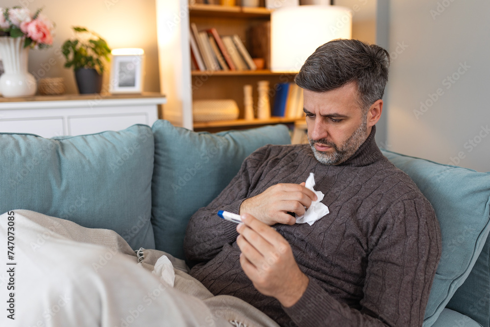 Man feeling sick lying in the bed and looking the thermometer. Sick man lying on sofa checking his temperature under a blanket at home in the living room Man feeling sick lying in the bed and looking the thermometer. Sick man lying on sofa checking his temperature under a blanket at home in the living room