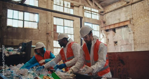 A trio of waste recycling plant workers in special protective uniforms and white helmets stand near the conveyor and lay out bottles depending on the color of the plastic at the plant photo