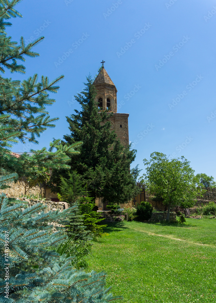 Monastery courtyard. Brick bell tower behind a coniferous tree. Earthen path to stone wall. Daylight, blue sky.
