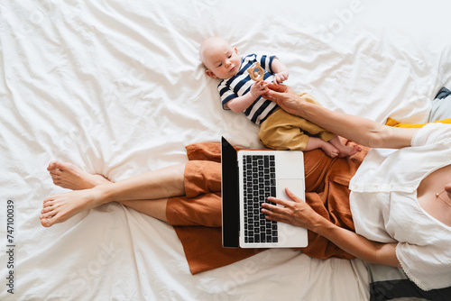 Mother working at laptop playing with baby in bed at home.