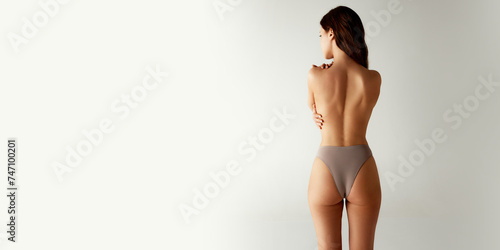 Rear view of half-naked woman in beige underwear standing hug herself against grey studio background. Body positivity. Concept of beauty treatments, dieting, female health, spa procedures. Ad