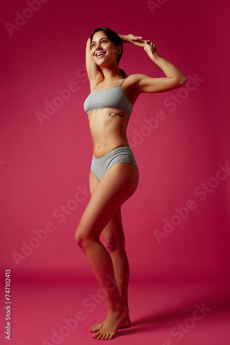 Full-length portrait of smiling woman posing in matching athletic wear holding hands behind her head against vivid pink studio background. Concept of diet, female health, femininity, plastic surgery.