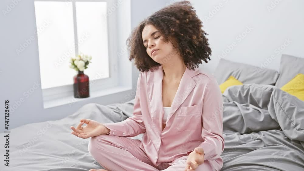 A young woman with curly hair practicing meditation in pajamas on a bed in a well-lit bedroom setting A young woman with curly hair practicing meditation in pajamas on a bed in a well-lit bedroom setting