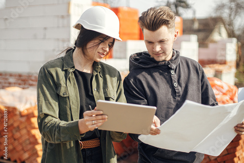 Stylish woman architect with tablet  and contractor man checking blueprints at construction site. Young engineer or construction workers in hardhat looking at plans of new modern house