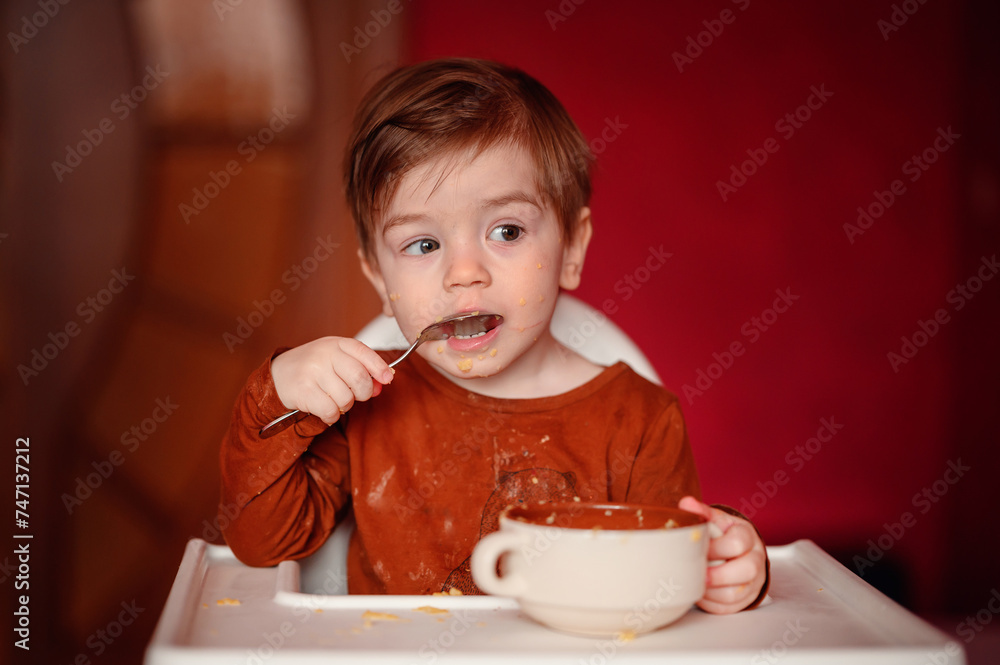 A young child focuses intently on scooping the remnants of a meal from a bowl, oblivious to the scattered food on the highchair tray and their own face. A young child focuses intently on scooping the remnants of a meal from a bowl, oblivious to the scattered food on the highchair tray and their own face.