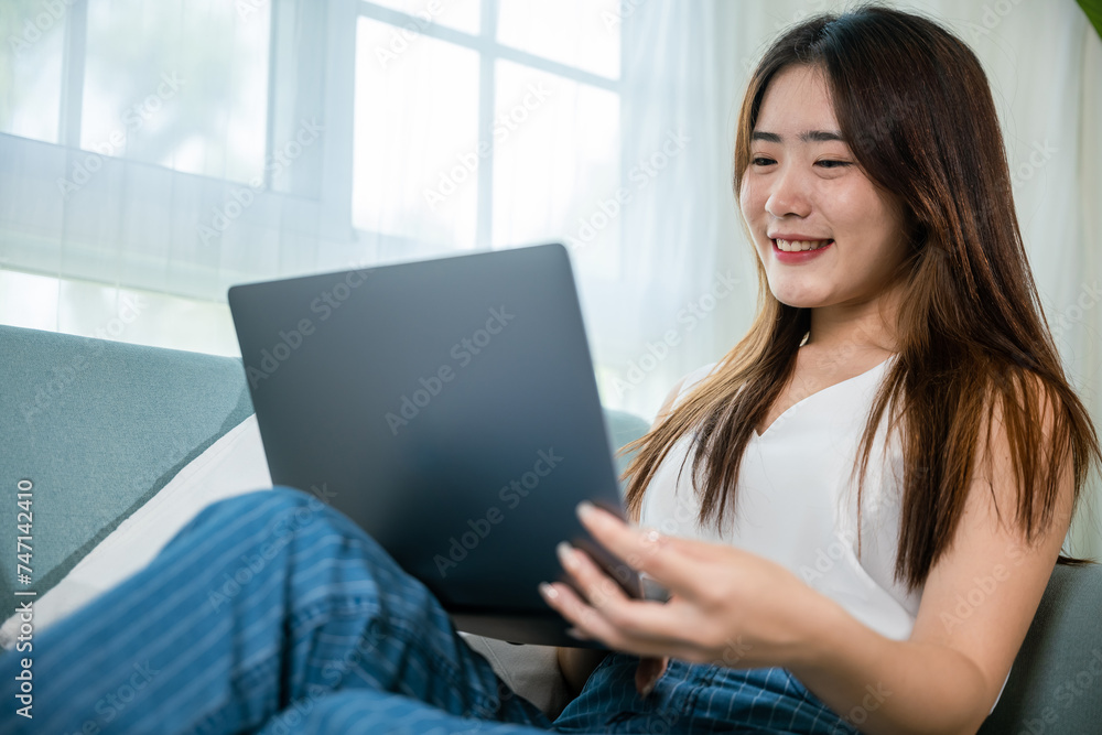 Happy woman typing email on notebook computer, Asian young female smiling sitting relaxing on sofa using laptop in living room at home, freelance browsing through the internet during free time Happy woman typing email on notebook computer, Asian young female smiling sitting relaxing on sofa using laptop in living room at home, freelance browsing through the internet during free time