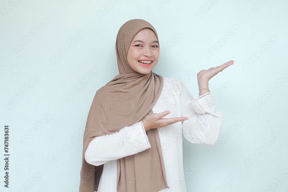 Young Asian Muslim woman wearing hijab, sweet smiling and see camera while pointing and showing to copy space beside her. Isolated in gray background. Young Asian Muslim woman wearing hijab, sweet smiling and see camera while pointing and showing to copy space beside her. Isolated in gray background.