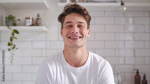 Smiling young man in white t-shirt standing in a white tiled bathroom with shelves and plants exuding a relaxed and happy vibe.