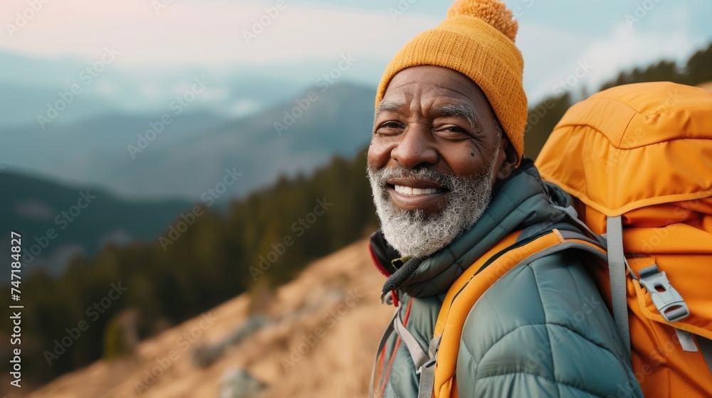 Naklejka premium An elderly man with a white beard and mustache wearing a blue jacket an orange beanie and carrying an orange backpack smiling and looking to the side standing on a mountain background.