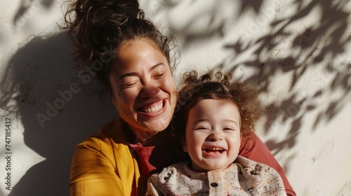A joyful mother and child sharing a moment of happiness with the mother's radiant smile and the child's innocent laughter set against a backdrop of a white wall with shadows from trees. photo