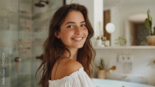 Smiling young woman with long brown hair wearing a white off-the-shoulder top standing in a modern bathroom with a white marble countertop and plants.