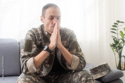 Portrait of calm serious Caucasian military man wearing camouflage uniform and cap sitting on a sofa, having depressed facial expression. © Angelov