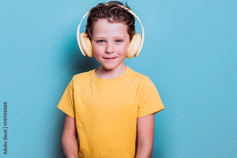 A cheerful school boy wearing a yellow t-shirt and headphones stands against a blue background, smiling warmly A cheerful school boy wearing a yellow t-shirt and headphones stands against a blue background, smiling warmly
