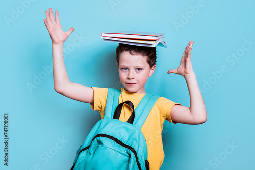 A school boy with a backpack strikes a playful pose, balancing a stack of books on his head against a light blue background photo