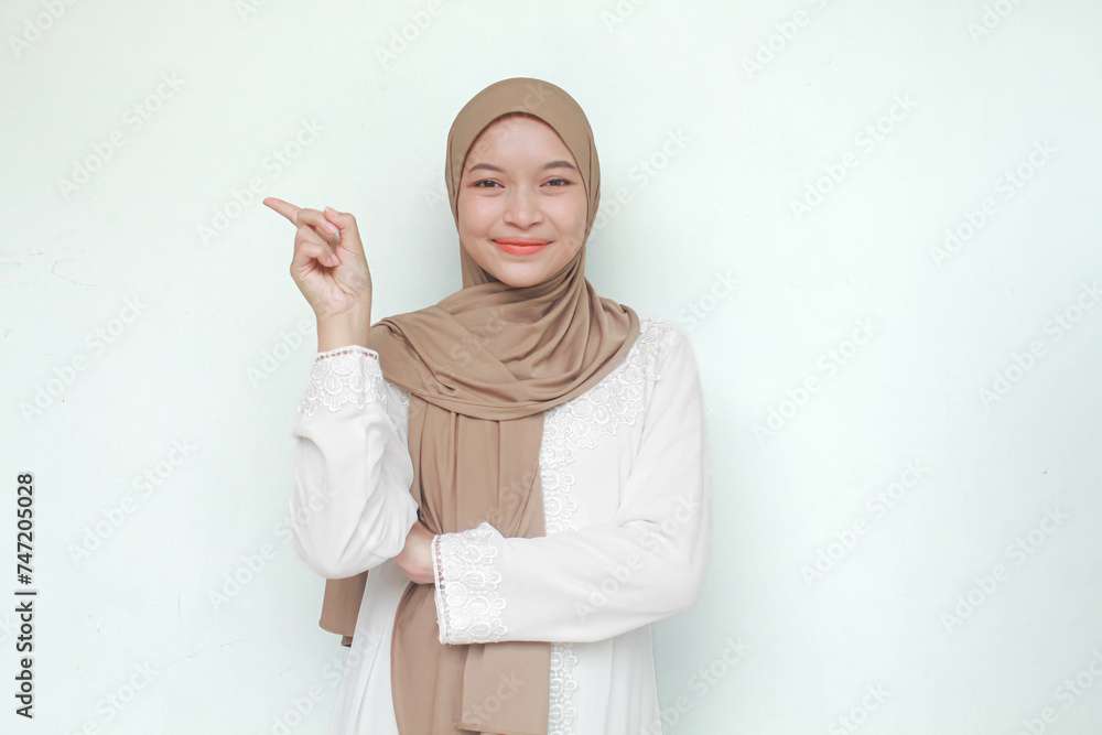 Young Asian Muslim woman wearing hijab, sweet smiling and see camera while pointing and showing to copy space beside her. Isolated in gray background. Young Asian Muslim woman wearing hijab, sweet smiling and see camera while pointing and showing to copy space beside her. Isolated in gray background.