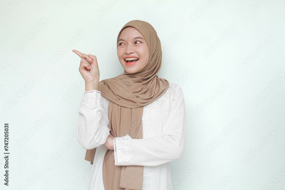 Young Asian Muslim woman wearing hijab, sweet smiling while pointing and showing to copy space beside her. Isolated in gray background. Young Asian Muslim woman wearing hijab, sweet smiling while pointing and showing to copy space beside her. Isolated in gray background.