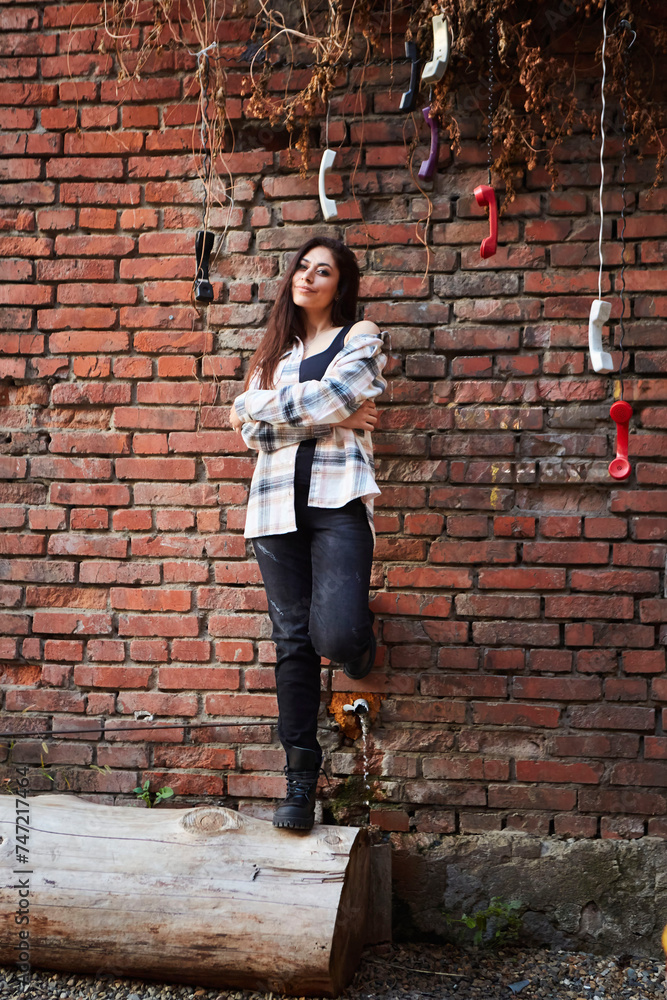 The girl is standing near a brick wall. Vintage handsets hang from the top. Women's clothing style is free urban The girl is standing near a brick wall. Vintage handsets hang from the top. Women's clothing style is free urban