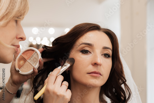 The girl makeup artist uses a brush to apply a highlighter to the light areas of the model s skin to give shine