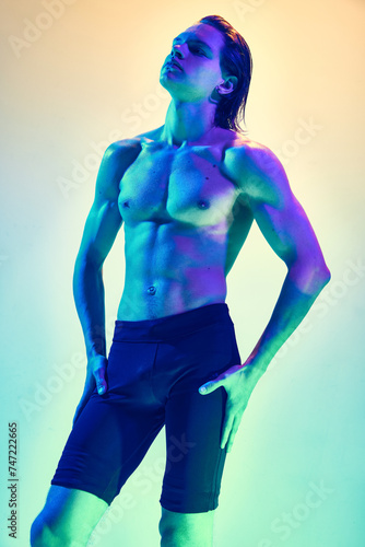 Confidence and beauty. Portrait of young man with muscular build body posing in blue-purple neon light against gradient studio background. Concept of natural beauty people, male health, masculinity.