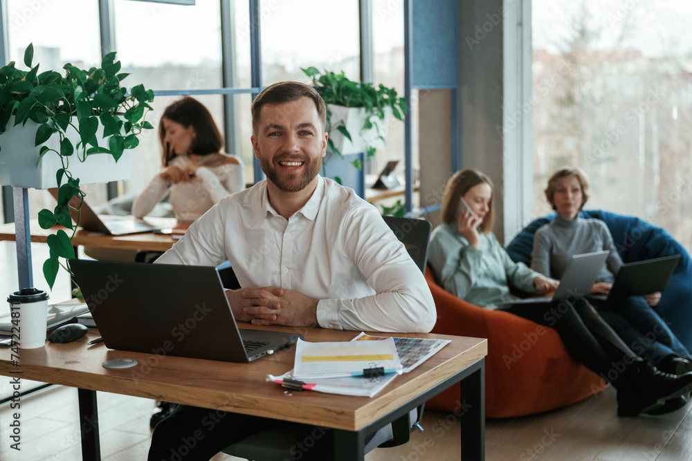 Cheerful friendly man is sitting by the table with laptop. People are working in the office with bean bags chairs in it Cheerful friendly man is sitting by the table with laptop. People are working in the office with bean bags chairs in it