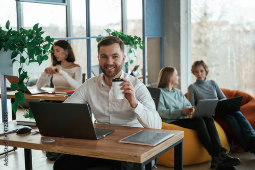 Man is smiling, sitting by the table with laptop. People are working in the office with bean bags chairs in it