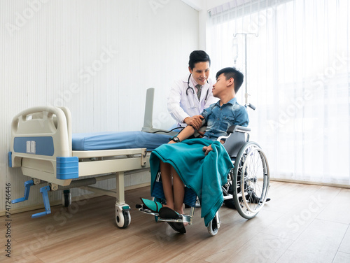 Compassionate Asian pediatric doctor listen and use measure pressure to a young handicapped child patient sitting on wheelchair beside a hospital bed with caring atmosphere in recovery room