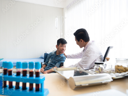 Asian caring pediatrician in a clinic interact with a smiling young disabled child boy sitting on wheelchair with medical equipment of blood test tube for examination in the foreground