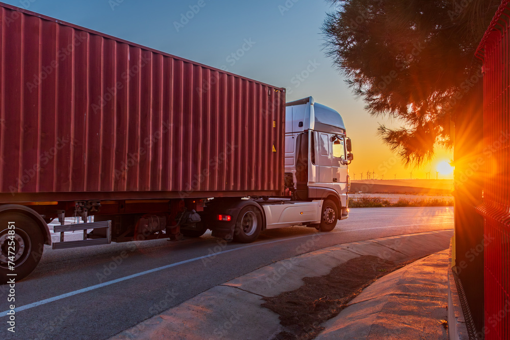 Truck with a semi-trailer with a maritime container driving on a conventional road and the evening sun in front. Side view.