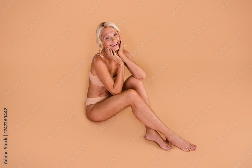 Full size portrait of pretty cheerful elderly person sit floor barefoot arms touch face isolated on beige color background Full size portrait of pretty cheerful elderly person sit floor barefoot arms touch face isolated on beige color background