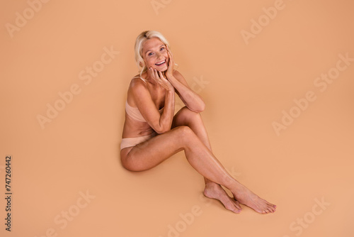 Full size portrait of pretty cheerful elderly person sit floor barefoot arms touch face isolated on beige color background