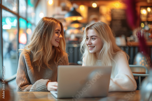 Two happy young women engaging in conversation while sitting in a cozy cafe, with a laptop on the table.