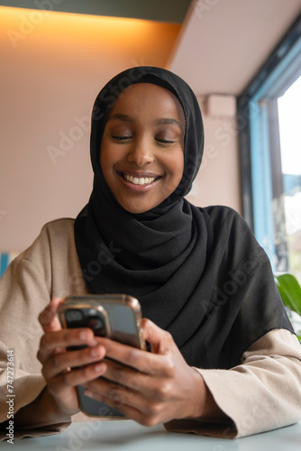 Smiling young woman in hijab using phone in cafe