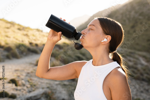 Rehydration. Female caucasian athlete drinking water from sporty bottle after tedious training jogging running. Water balance with electrolytes. Yoga outdoors in the mountains