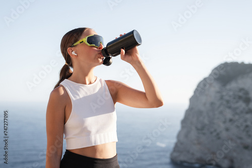 Water balance with electrolytes. Young sportive female athlete in fitness outfit drinking water after training yoga sportive exercises. Physical work out outdoors by the sea, in the mountains