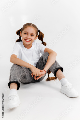 Stylish smiling kid girl model wearing jeans and white t-shirt sitting and posing on white studio background