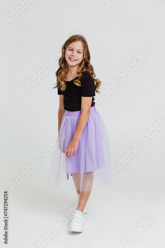 Smiling preteen girl in black t-shirt, violet skirt and white sneakers dancing and having fun on white background