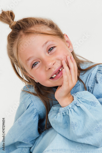 Portrait of cute toothless kid girl in blue dress arm touch chin on white studio background. Soft focus