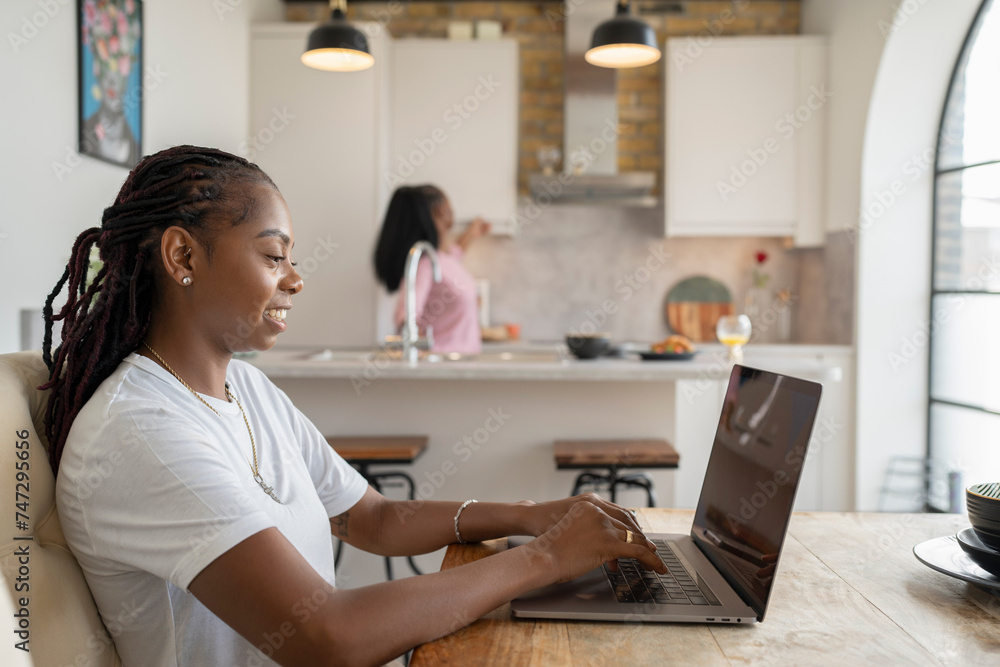Woman working on laptop while girlfriend preparing breakfast in kitchen - obrazy, fototapety, plakaty 