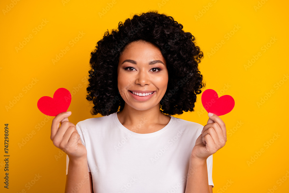 Portrait of lovely nice girl with curly hairstyle wear white t-shirt hold two red paper hearts isolated on vivid yellow color background Portrait of lovely nice girl with curly hairstyle wear white t-shirt hold two red paper hearts isolated on vivid yellow color background