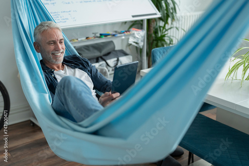 Smiling mature man lying in hammock and using digital tablet