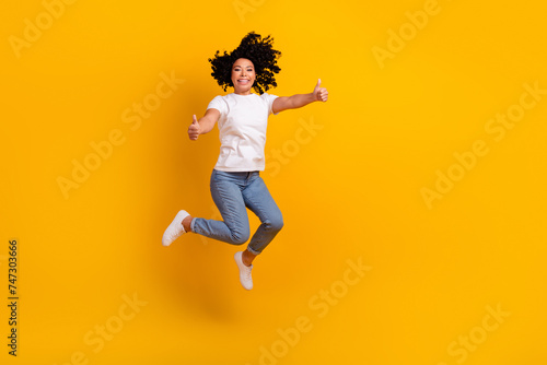 Full length photo of confident pretty lady dressed white t-shirt showing thumbs up jumping high empty space isolated yellow color background
