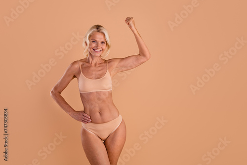 Photo of excited confident middle age woman wear bikini rising arm showing biceps empty space isolated beige color background