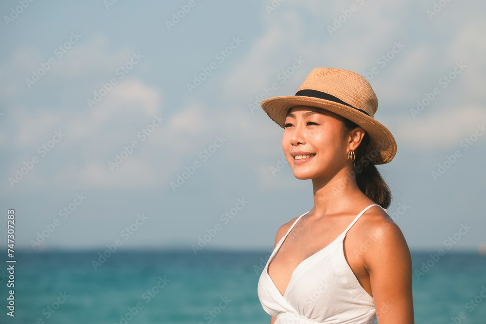 Portrait of smiling young Asian woman wearing a straw hat standing at the beach on vacation.