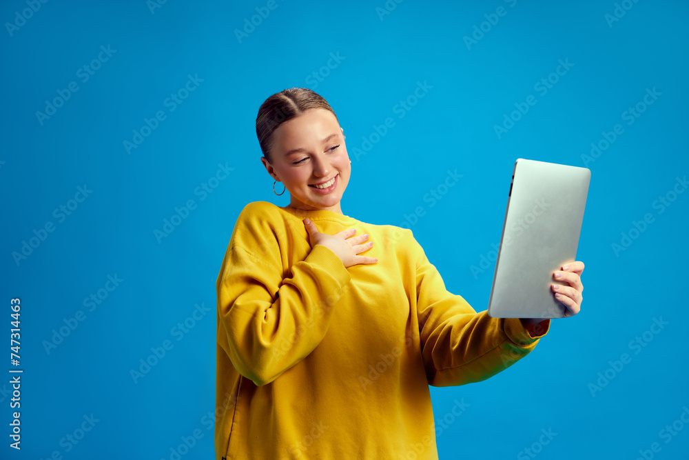 Young girl, student in yellow sweatshirt holding tablet and having online video call, lesson against blue studio background. Concept of youth, human emotions, casual fashion Young girl, student in yellow sweatshirt holding tablet and having online video call, lesson against blue studio background. Concept of youth, human emotions, casual fashion