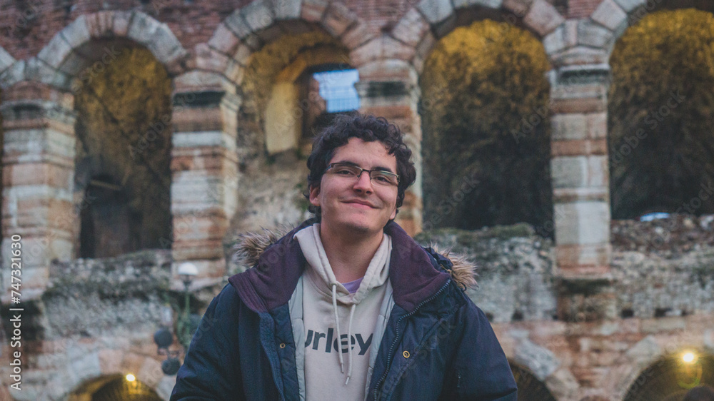 White young man with glasses and curly hair smiling to the camera with the Verona Arena in the background