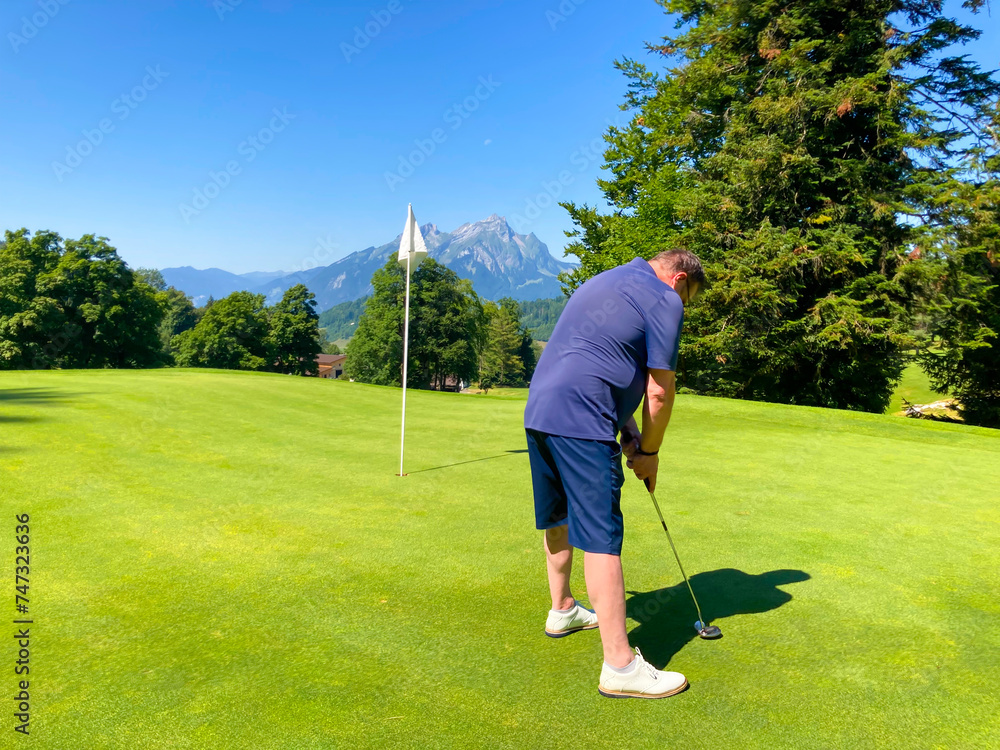 Fototapeta premium Golfer on Putting Green with Mountain Pilatus and the Moon View in a Sunny Summer Day in Bürgenstock, Nidwalden, Switzerland.