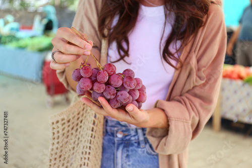 Cropped shot of a young woman with a net bag picking fresh grapes on farmers market. Shopping for organic local produce fruits. Close up, copy space, background.